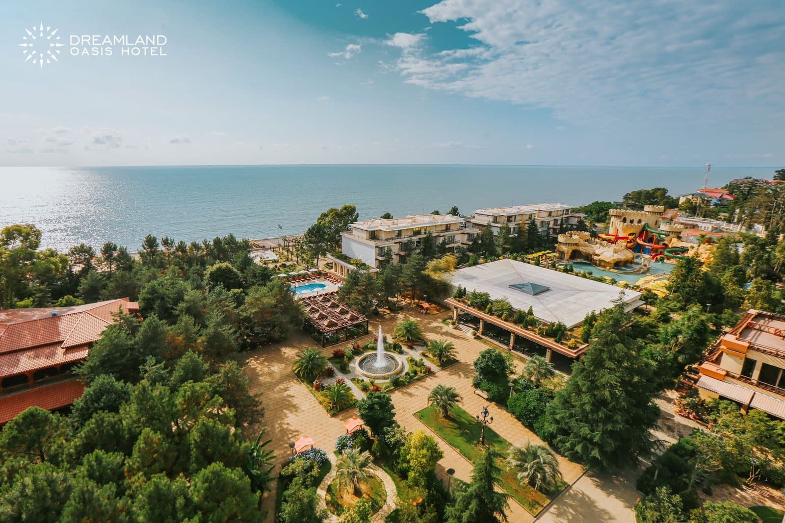 Панорама курорта сверху: парк с фонтаном, корпуса под красными и белыми крышами, вдали горки аквапарка и море