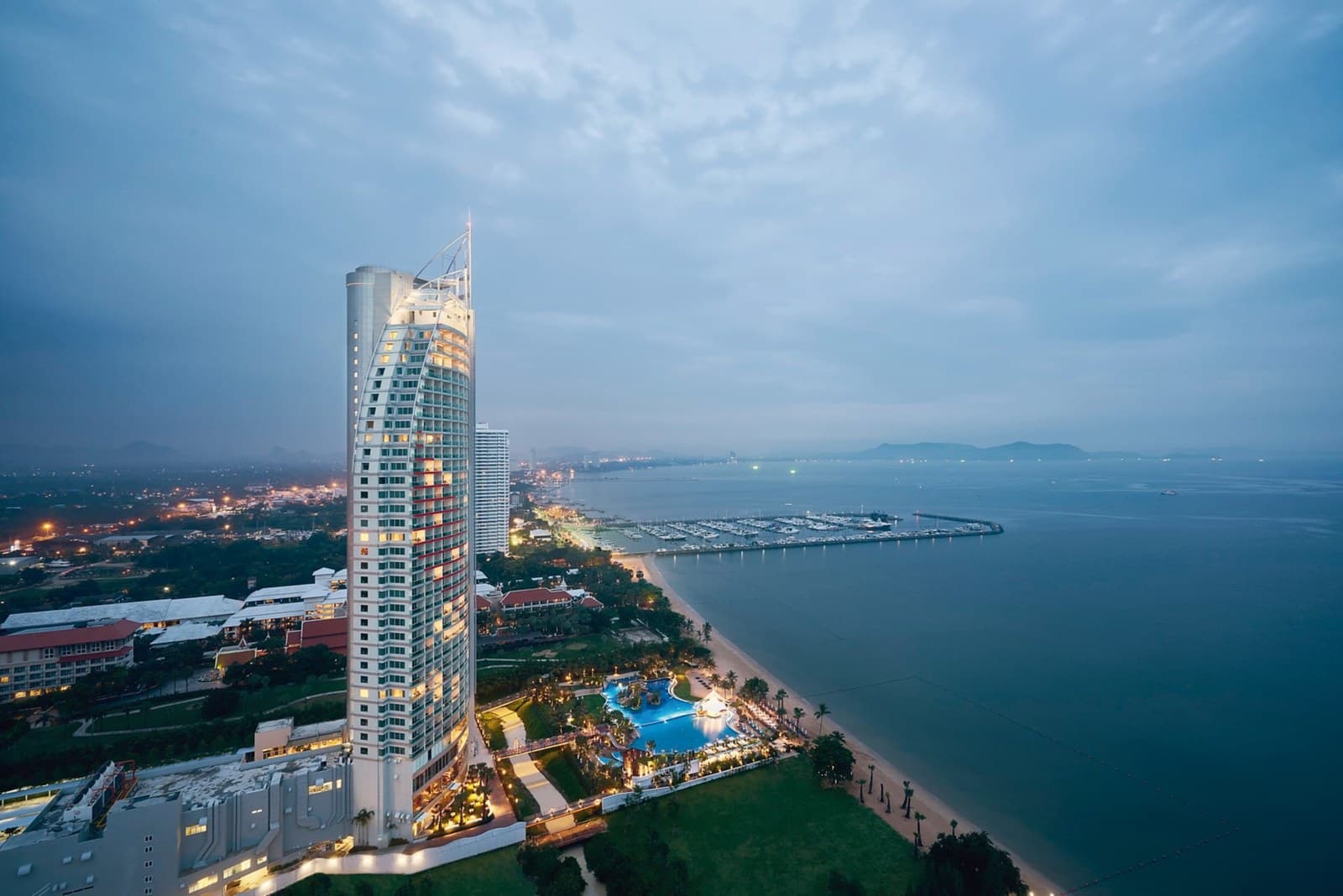 Высотная башня отеля Mövenpick Siam на берегу залива в сумерках, внизу подсвеченный бассейн-лагуна и линия пляжа