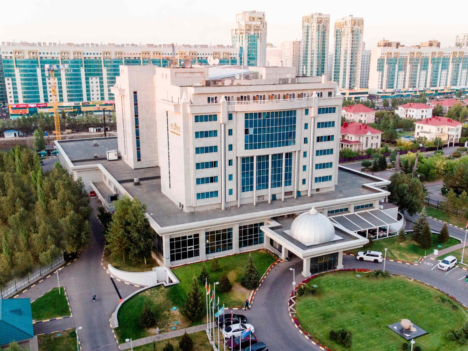 Здание отеля Rixos President Astana с высоты: светлый корпус с синими окнами, купол над входом, круговой подъезд с газоном, на заднем плане жилые дома