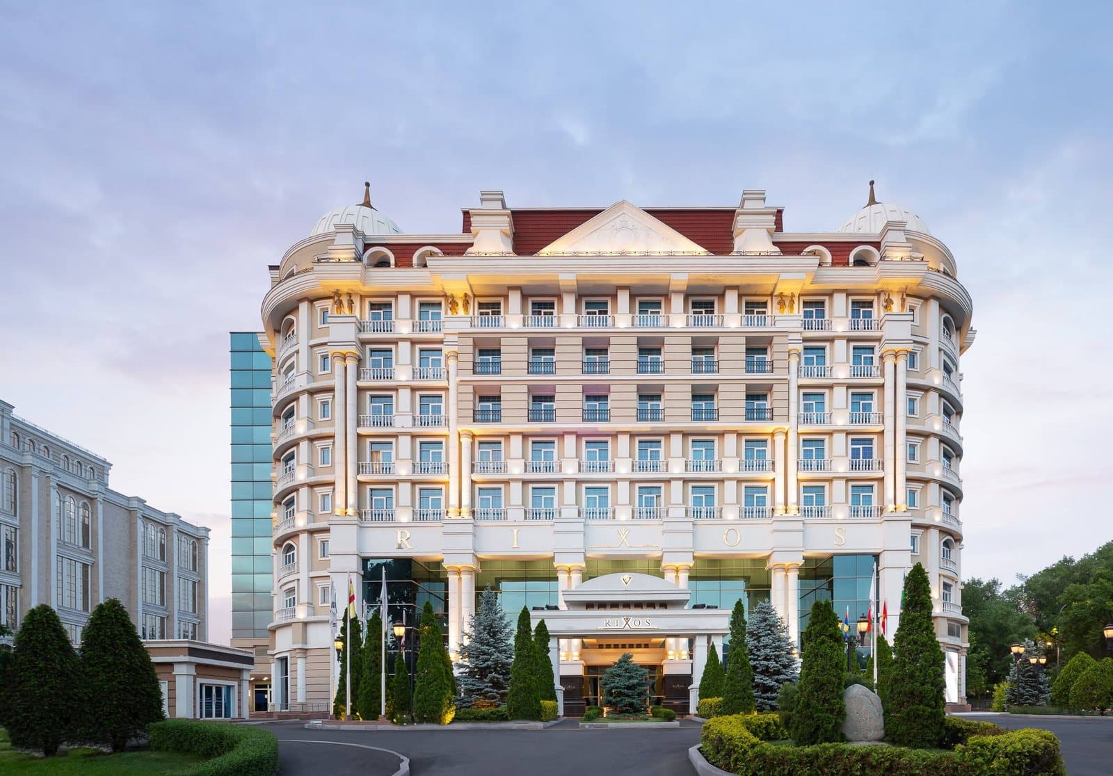 Главный фасад отеля Rixos Almaty в сумерках: светлое здание с колоннами, балконами и красной крышей, перед входом ели и клумбы