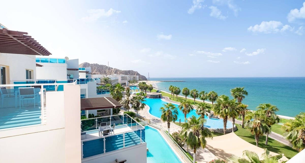 Вид с террасы на бассейн среди пальм, газоны и пляж Radisson Blu Resort, Fujairah у гор