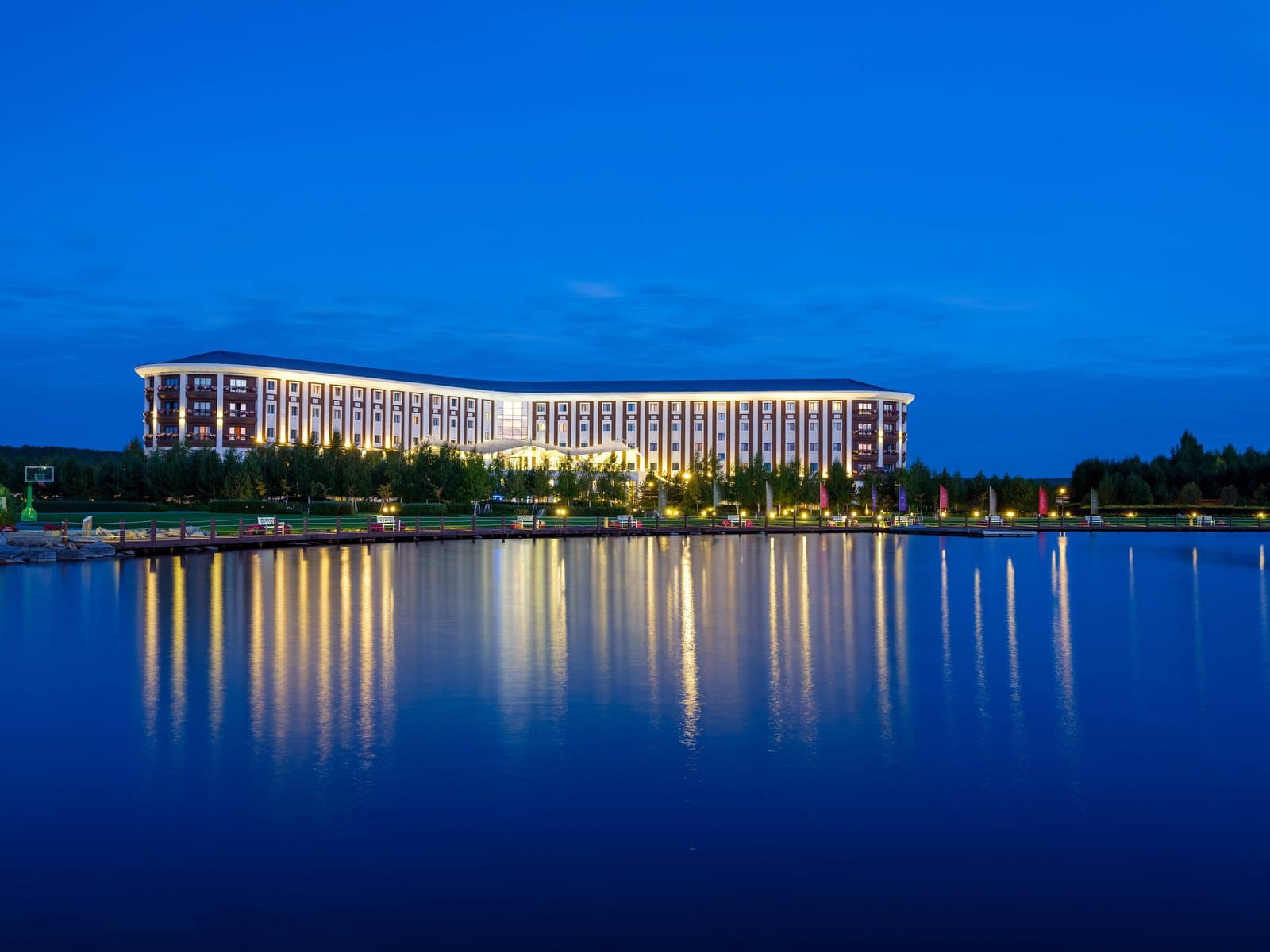 Корпус отеля Rixos Borovoe вечером: длинное здание в тёплой подсветке на берегу, огни отражаются в тёмно-синей воде
